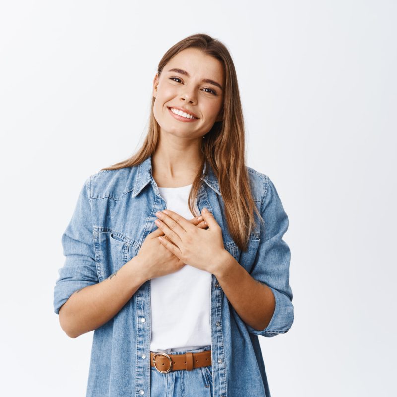 Portrait of touched young woman feeling flattered, holding hands on heart and smiling white teeth, say thank you, appreciate help, being grateful for nice gesture, white background.